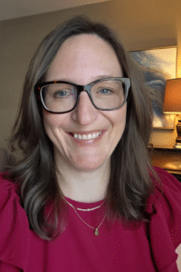 Smiling headshot of a Caucasian woman with brown, straight hair and glasses wearing a maroon top