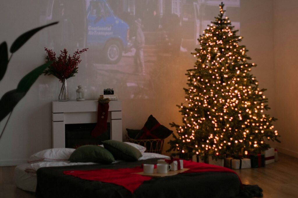 Empty, warmly lit living room with lit Christmas tree (white lights), fireplace with flowers on mantle, and Christmas decor