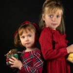 lukas-parnican-sxscElnGXb4-unsplash - Bridging Divorce Solutions Two young girls with serious facial expressions standing back to back wearing Holiday-themed dresses and holding small packages in their hands