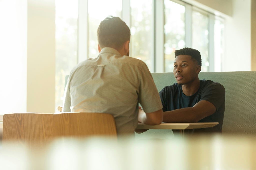 Two men sitting and talking at a table next to a window