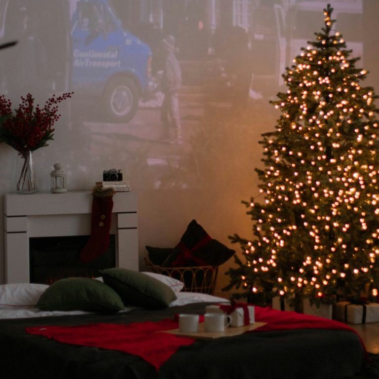 Empty, warmly lit living room with lit Christmas tree (white lights), fireplace with flowers on mantle, and Christmas decor