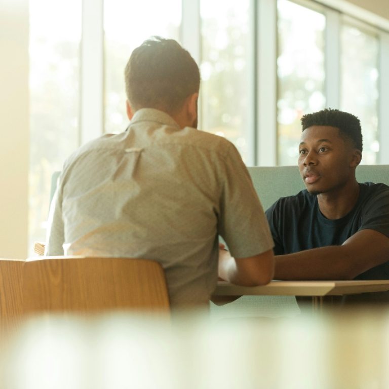 Two men sitting and talking at a table next to a window