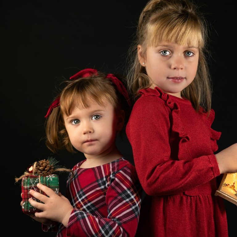 Two young girls with serious facial expressions standing back to back wearing Holiday-themed dresses and holding small packages in their hands